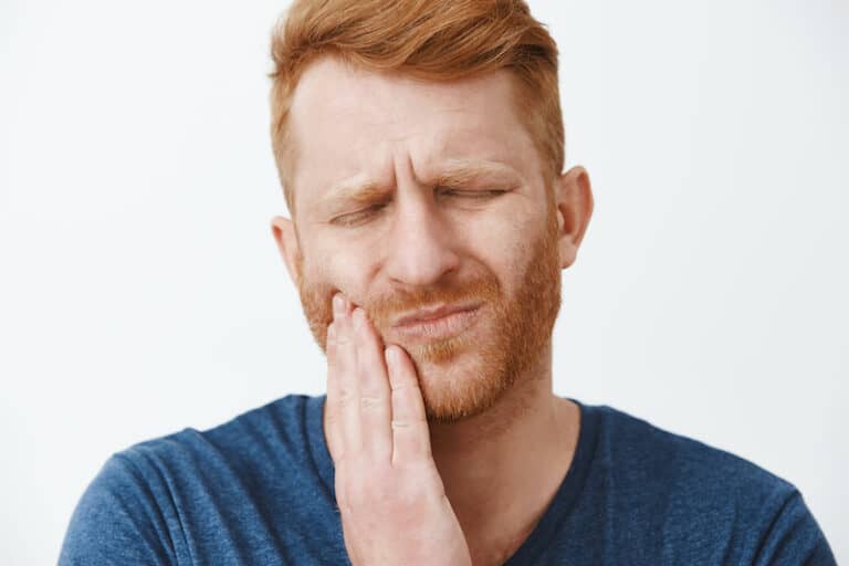 Close-up shot of redhead man with beard feeling pain in teeth, frowning and making suffering expression with closed eyes, touching cheek, having need to call dentist for curing decay or rotten tooth